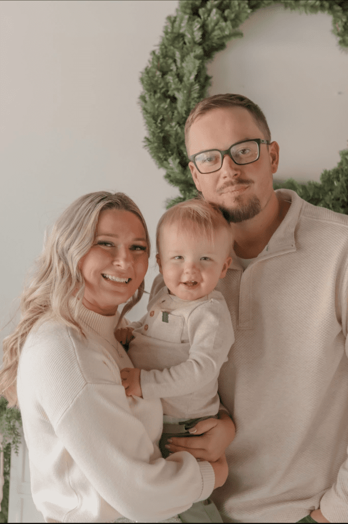 Family portrait with a woman, man, and toddler smiling, dressed in cozy sweaters, against a backdrop of greenery, representing community and family values associated with PMT Lawn & Landscape.